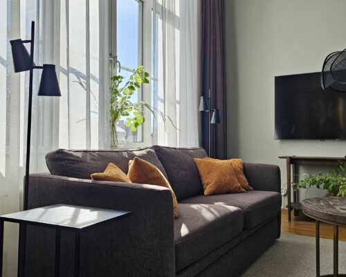 Bilden föreställer en soffa med soffbord, sidobord, prydnadskuddar och ett stort fönster. Rummet är sviten på Hotel Skansen Öland