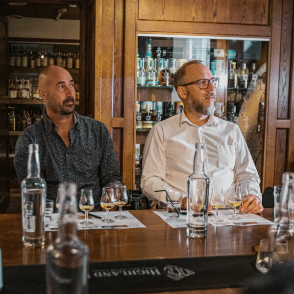 Två män sitter vid ett bord under en whiskyprovning på Hotel Skansen, omgivna av olika whiskysorter. De ser intresserade ut, vilket skapar en atmosfär av lärande och njutning för whiskyentusiaster.