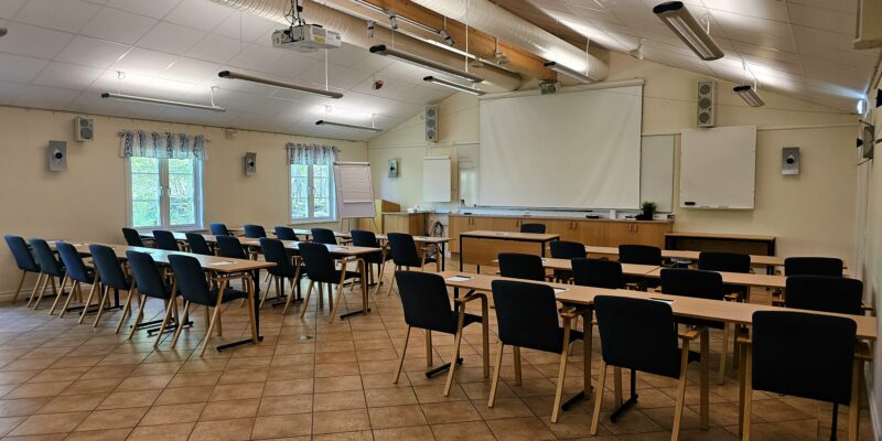 På bilden syns konferenslokalen kallad Tingshussalen hos Hotel Skansen på Öland. Lokalen är möblerad med 3 rader av stolar och bord för 30 personer, blädderblock och en vit duk till en projektor.