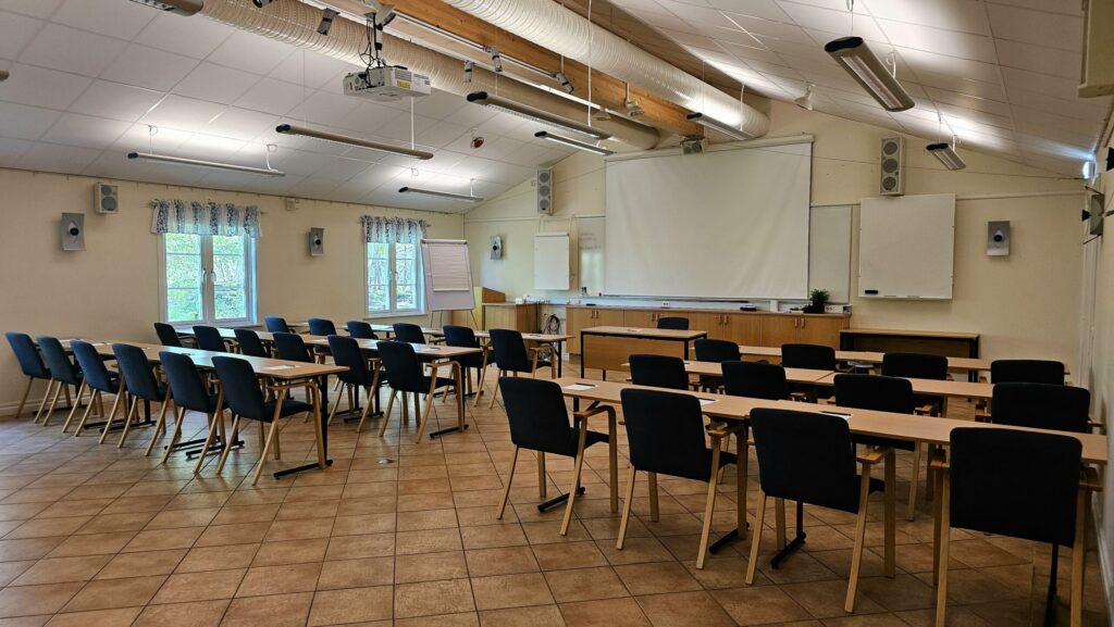 På bilden syns konferenslokalen kallad Tingshussalen hos Hotel Skansen på Öland. Lokalen är möblerad med 3 rader av stolar och bord för 30 personer, blädderblock och en vit duk till en projektor.