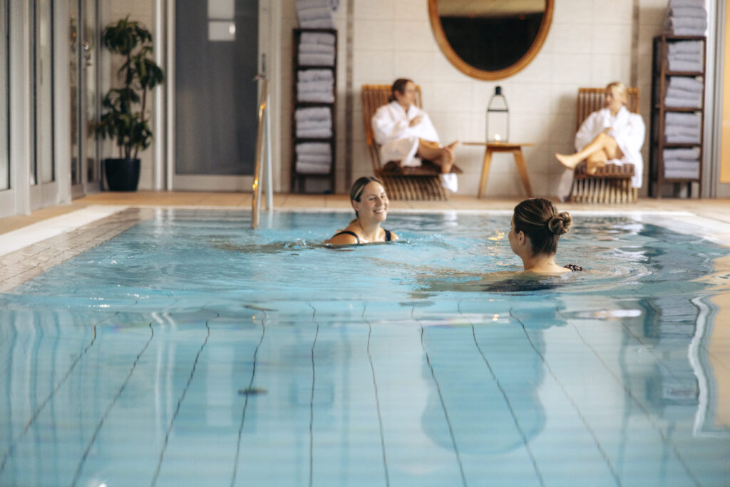 Två personer njuter av ett rogivande bad i Hotel Skansens inomhuspool, medan två andra kopplar av i loungestolar i bakgrunden, omgivna av vikta handdukar och mjuk belysning.