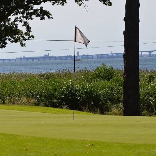 Vy över en grön golfbana på Hotel Skansen med flagga i förgrunden och havet samt Ölandsbron i bakgrunden. En avkopplande miljö där natur och sport möts.