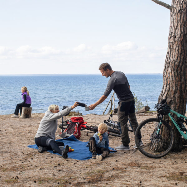 En grupp vid havet där en man räcker över en termos till en sittande kvinna – en aktiv paus i naturen nära Hotel Skansen.
