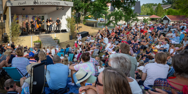 En stor publik sitter på gräset och lyssnar på livemusik från scenen under ett sommarevenemang på Hotel Skansen.