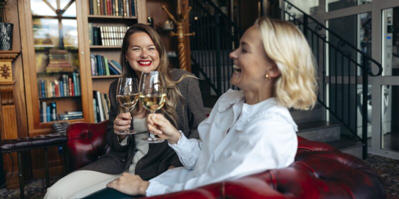 Två kvinnor skålar med vin i hotellbibliotekets mysiga miljö med bokhyllor och röd soffa på Hotel Skansen.