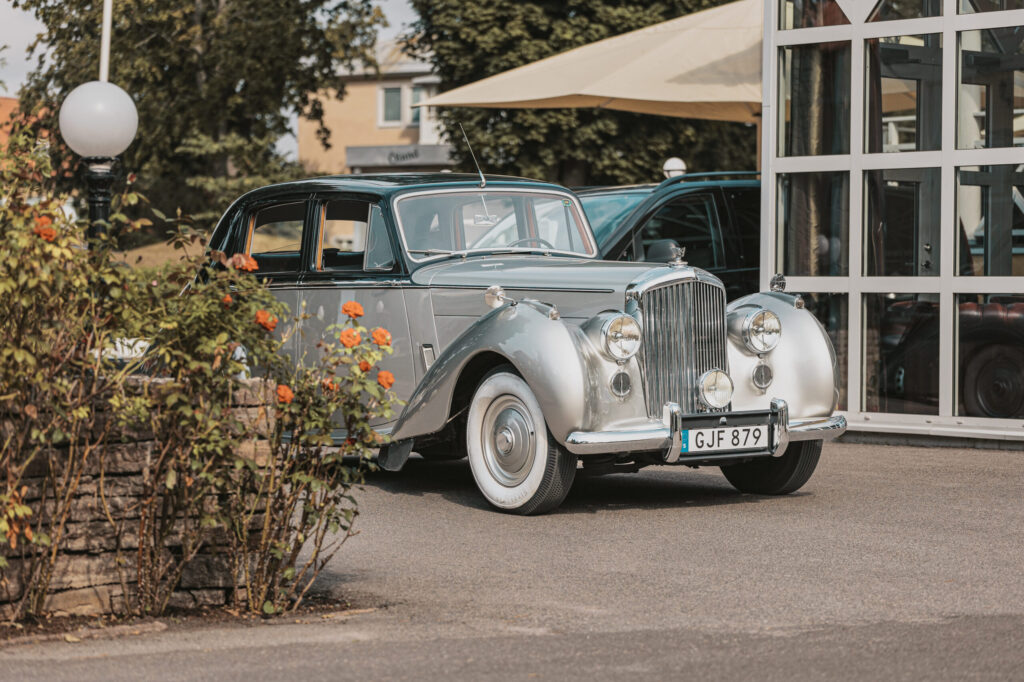 På bilden syns en bil som står mellan en stenmur klädd med en buske och fönstrena till hotel Skansen.