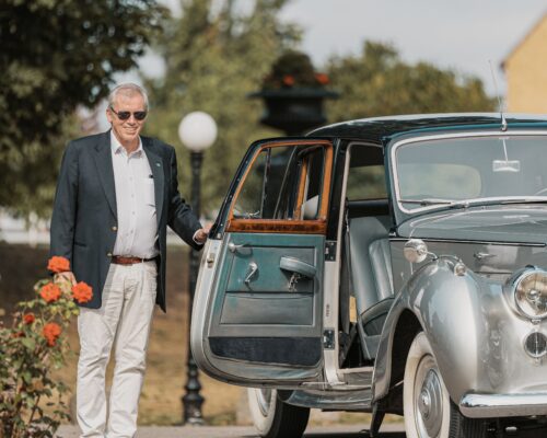 På bilden syns en silverfärgad bil utanför Hotel Skansen. Bilens ena passagerardörr hålls upp av en man klädd i kostym.