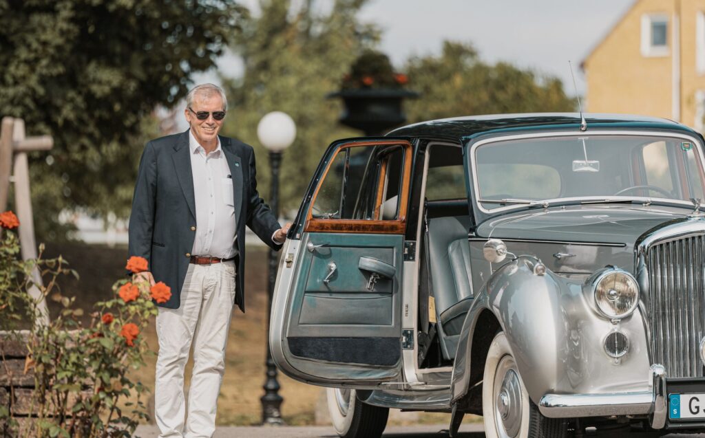 På bilden syns en silverfärgad bil utanför Hotel Skansen. Bilens ena passagerardörr hålls upp av en man klädd i kostym.