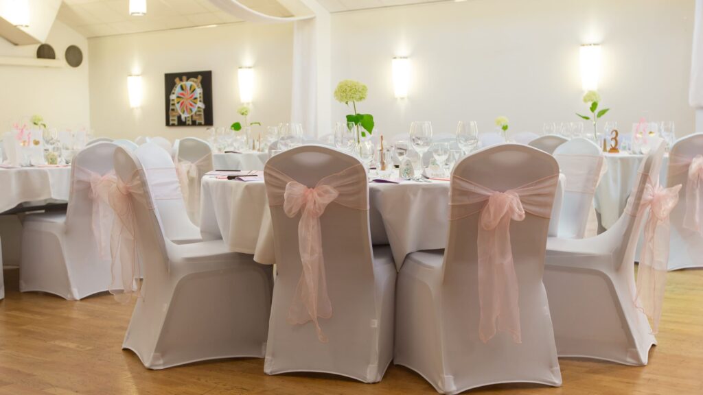 Bakifrån vy över flera dukade bord med vita dukar och stolsöverdrag med rosa rosetter på Hotel Skansen.