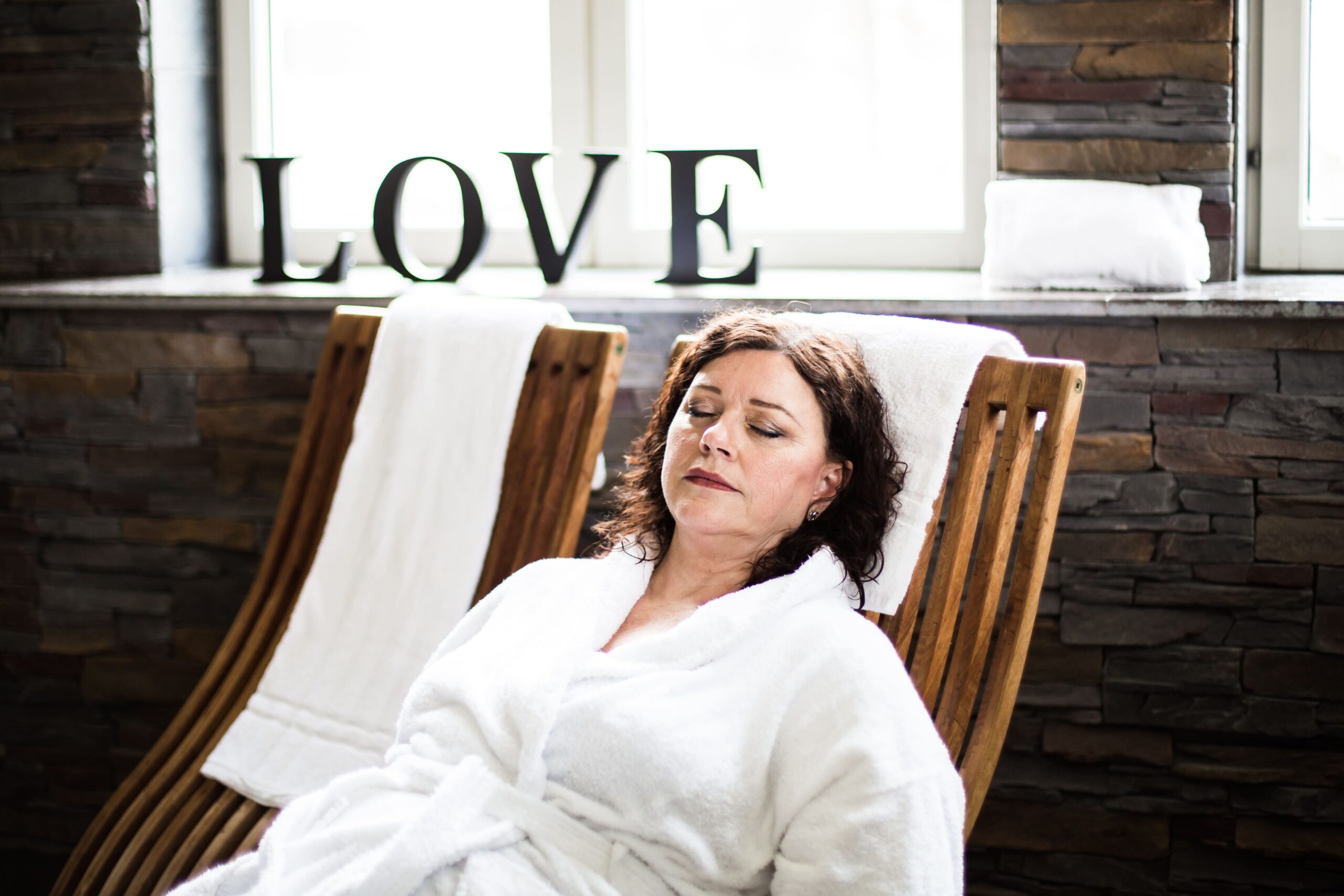 En kvinna vilar med slutna ögon i en vilstol iklädd morgonrock på Hotel Skansens spa, med ordet "LOVE" i bakgrunden – en stund av inre ro.