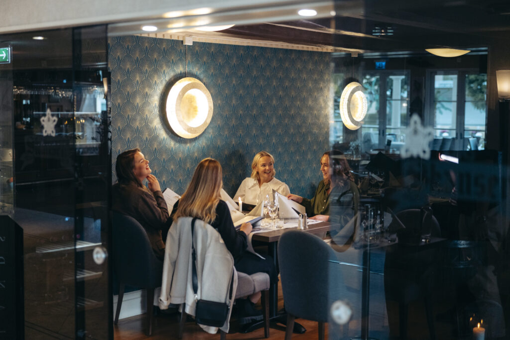 Fyra kvinnor i varmt samtal runt ett dukat bord i Hotel Skansens stämningsfulla restaurangmiljö, omgivna av mjukt ljus och mönstrade väggar.
