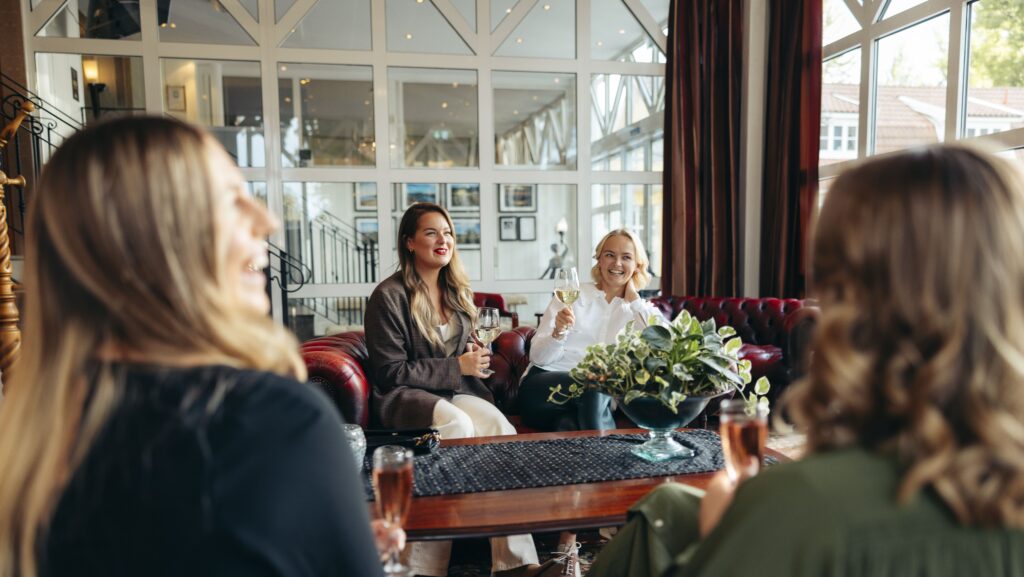 En grupp vänner sitter i en elegant lounge på Hotel Skansen, skrattar och njuter av vita vinglas. I förgrunden står en grön växt på bordet, vilket skapar en inbjudande och avslappnad atmosfär för social samvaro.