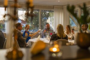 En varm kväll på Hotel Skansen där ett sällskap skålar runt ett elegant dukat bord i mjukt stearinljus, med leenden och avslappnad stämning i ett ljust inglasat rum.