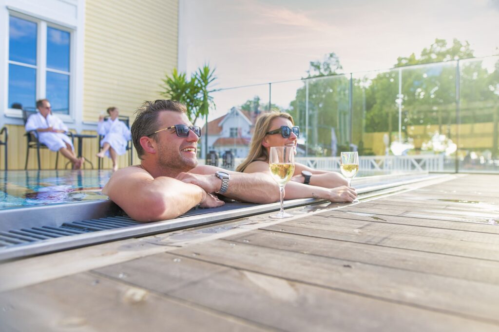Ett leende par lutar sig mot poolkanten med varsitt glas vitt vin, medan eftermiddagssolen lyser över Hotel Skansens utomhusspa