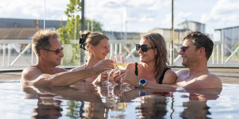 Fyra personer skålar glatt med bubbel i utomhuspoolen i solsken på Hotel Skansen.