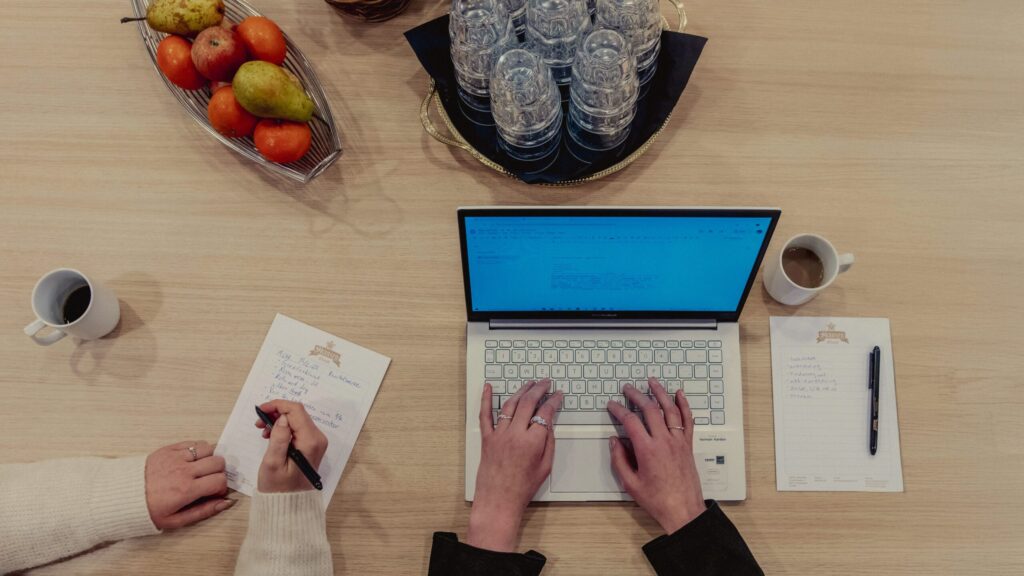 Ett fågelperspektiv över ett mötesbord med laptop, anteckningar, kaffe och frukt – en energigivande paus i Hotel Skansens konferensmiljö.