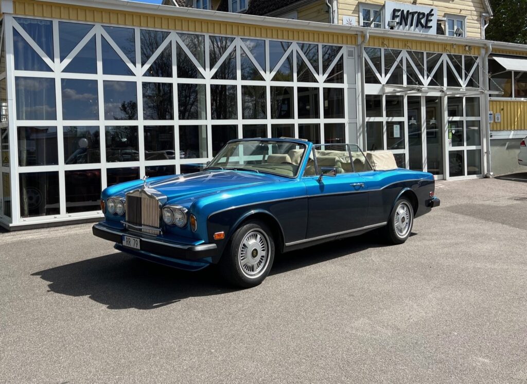 I bakgrunden på bilden syns Hotel Skansen fasad som är glastäckt. Framför fasaden står en blå Rolls Royce parkerad med nedcabbat tak.