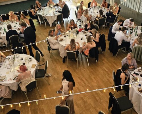 Bild tagen uppifrån på en bal i Hotel Skansens festlokal. Man ser flera runda bord med balgäster som sitter och äter. Över borden hänger ljusslingor