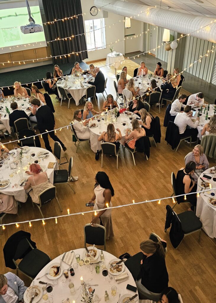 Bild tagen uppifrån på en bal i Hotel Skansens festlokal. Man ser flera runda bord med balgäster som sitter och äter. Över borden hänger ljusslingor