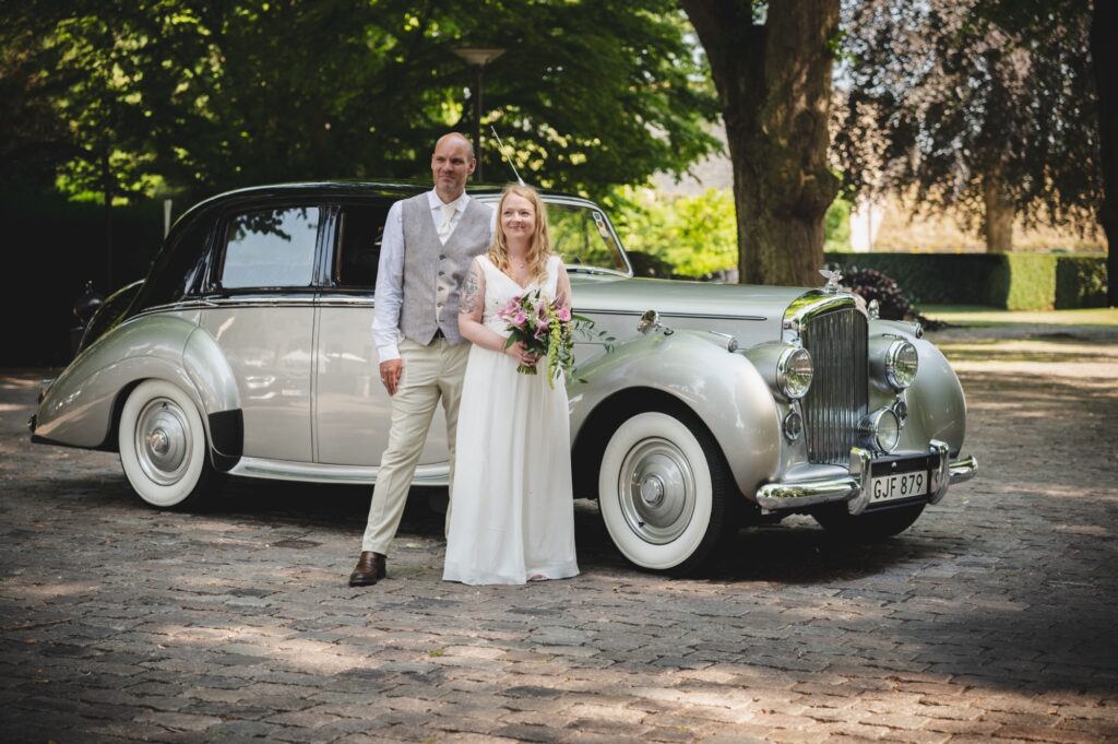 Ett brudpar står tillsammans och ler mot kameran fram Hotel Skansens ena bröllopsbil, en bentley. Mannen är klädd i kostymbyxor, skjorta och väst, kvinnan i brudklänning och håller en brudbukett i handen.