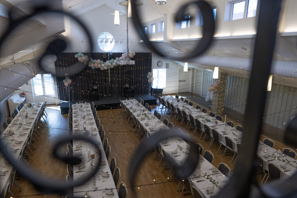 Bild ovanifrån på Gästgifvarsalen hos Hotel Skansen. På bilden syns fyra stycken långbord uppdukade med heltäcktande dukar, servetter, bestick, glas, murgröna och blommor. Lokalen är pyntad med ljusslingor över borden och ballonger utplacerade på flertalet ställen.
