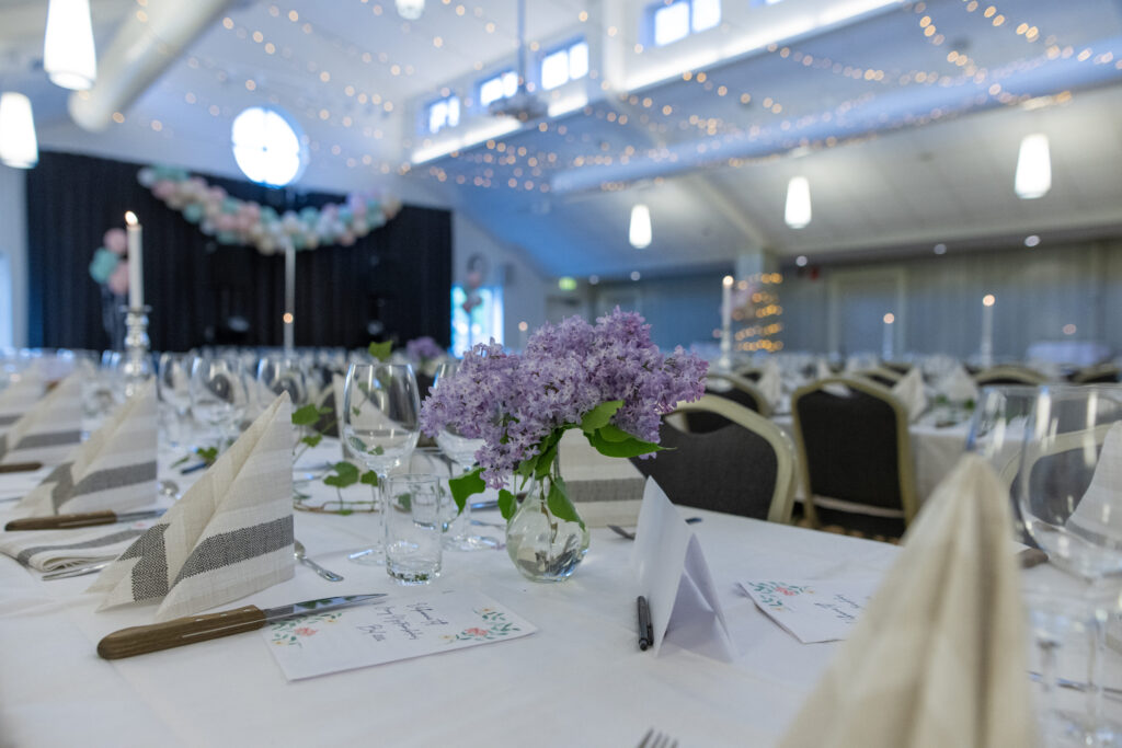 Bilden föreställer en närbild av dukning för en student bal hos Hotel Skansen med bestick, glas, blommor, servetter. I bakgrunden ser man att lokalen är pyntad med ljusslingor och ballonger.
