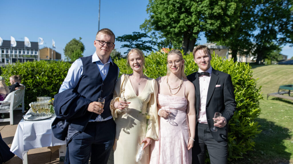 Två par uppklädda i balkläder med varsitt glas mousserande i handen ler mot kameran i parken på Hotel Skansen